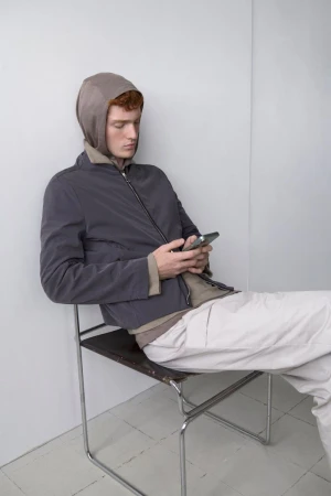 Model Jesse Sachet-Dufraisse seated on a metal frame chair looking at a phone in a grey zip jacket and off-white trousers.