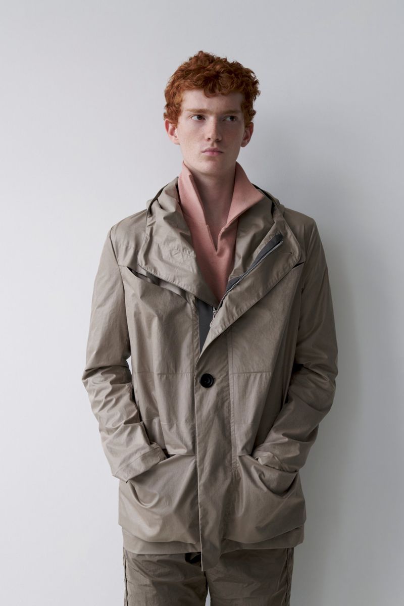 A red-haired male model poses in a light brown jacket and matching pants against a plain white backdrop in a studio fashion shot.