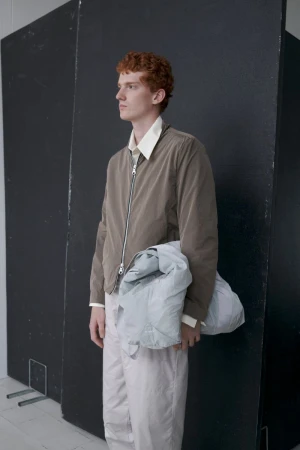 A red-haired male model poses indoors wearing a light brown zip-up jacket, white collared shirt, light trousers, and carrying a blue-grey jacket.