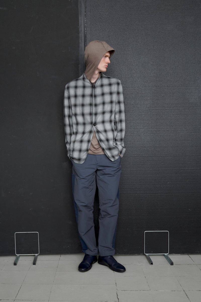 Full-length shot of a male model in a hooded, plaid blazer and pants, standing against a dark wall with grey tiled flooring.