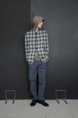 Full-length shot of a male model in a hooded, plaid blazer and pants, standing against a dark wall with grey tiled flooring.