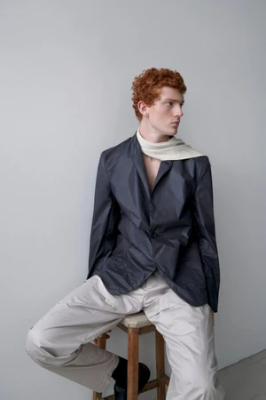 Model Jesse Sachet-Dufraisse seated on a wooden stool wearing a dark nylon blazer and light trousers against a neutral backdrop.