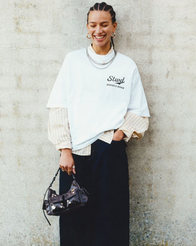 Smiling woman in a white graphic tee and long black skirt carries a purse in front of a textured concrete wall.