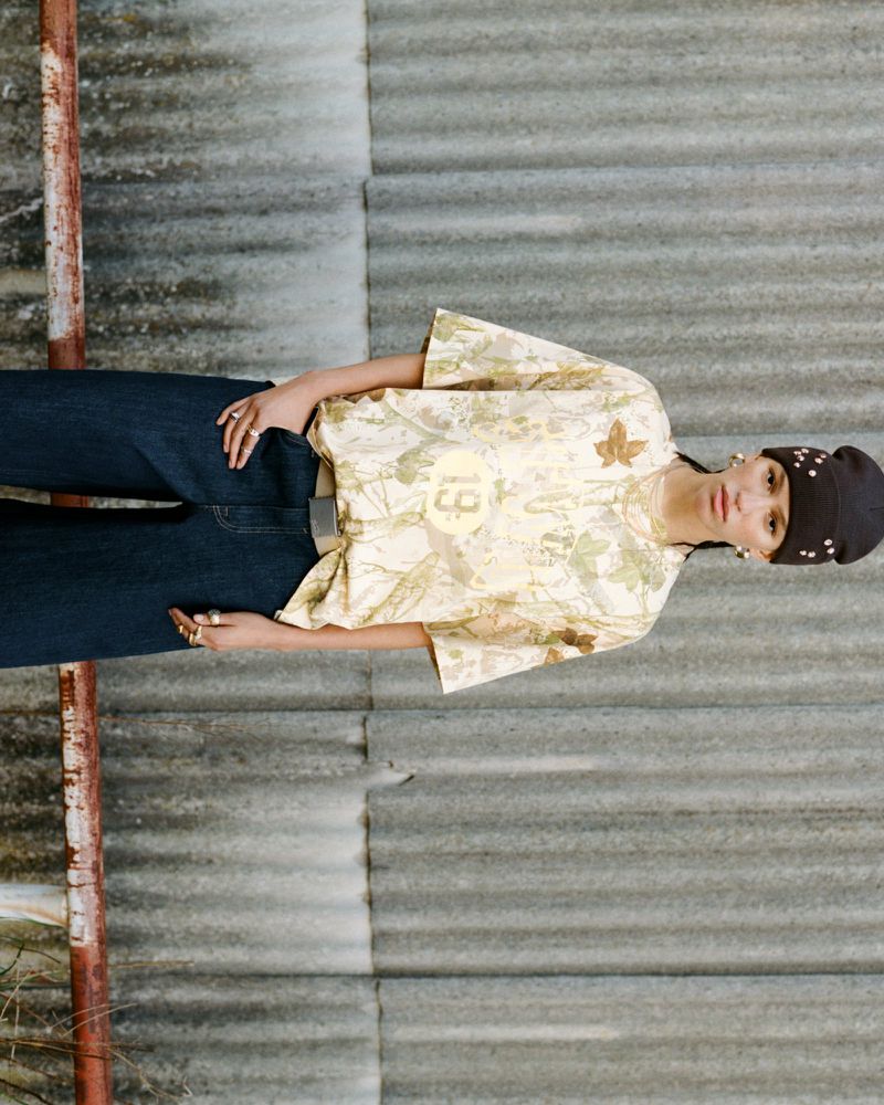 Adult female with a neutral expression wearing a patterned t-shirt and dark beanie poses outdoors against a corrugated wall.
