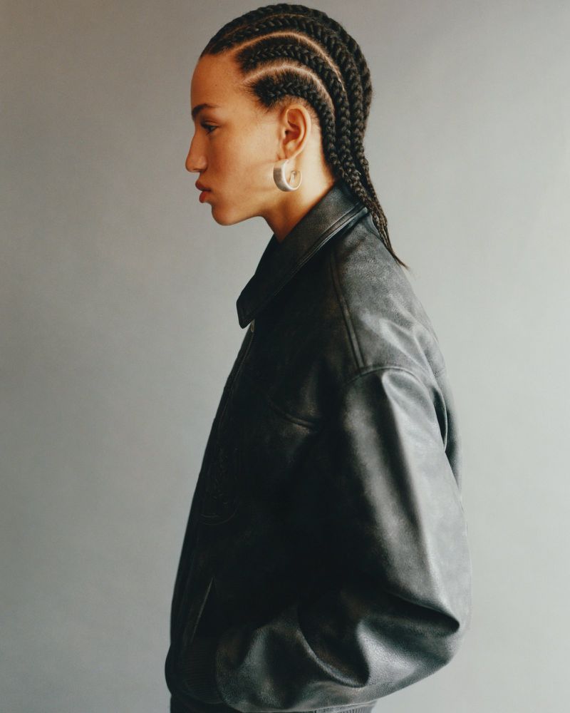 Side profile of a young adult woman with cornrow braids wearing a black leather jacket against a grey backdrop in a studio setting.