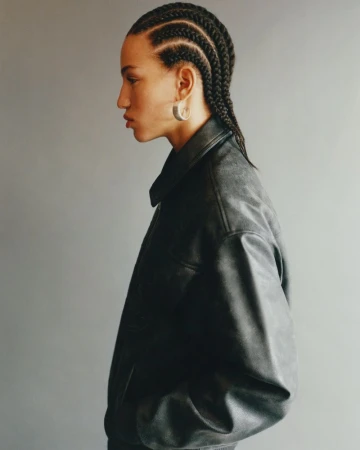 Side profile of a young adult woman with cornrow braids wearing a black leather jacket against a grey backdrop in a studio setting.