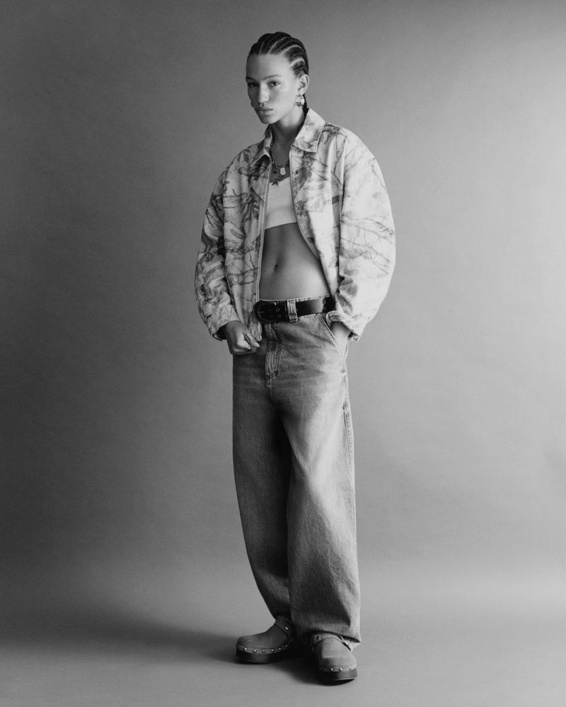 A fashion-forward woman poses in a light jacket, bralette, and baggy jeans against a neutral backdrop, showcasing contemporary style and design.