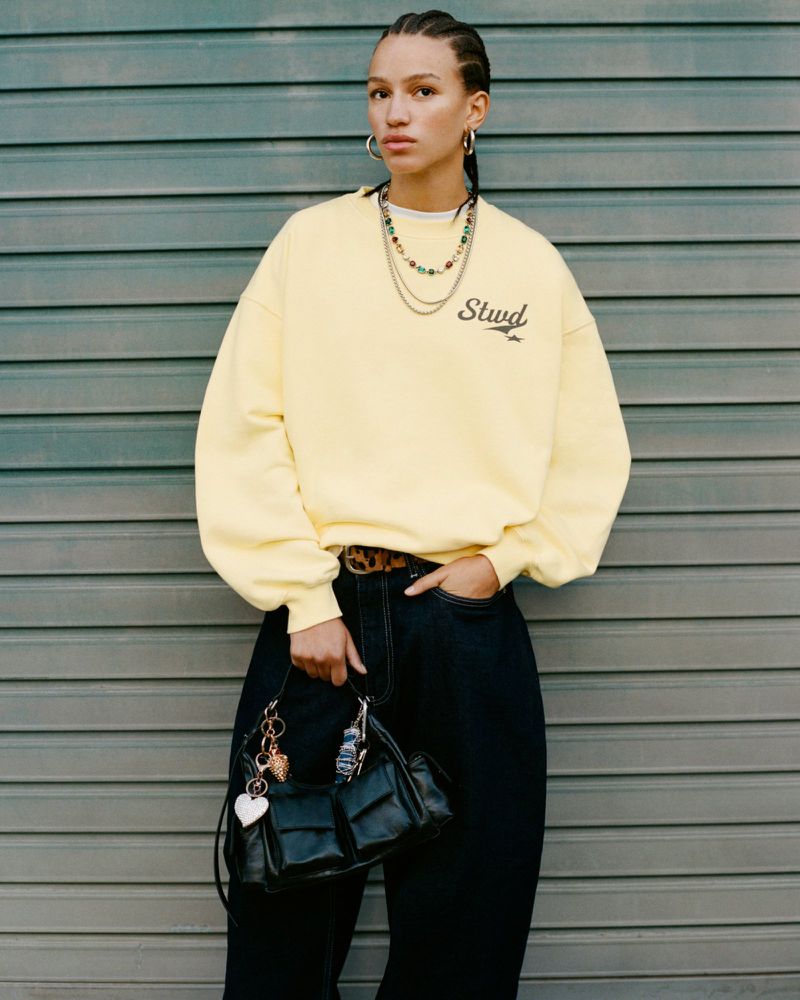 A woman poses against a corrugated metal background, showcasing a yellow sweatshirt, dark denim, and a structured black leather handbag.