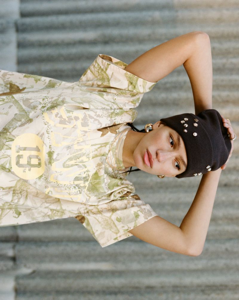 Woman with a black beanie poses with her arms up in an outdoor environment wearing an off-white, patterned short-sleeve shirt.