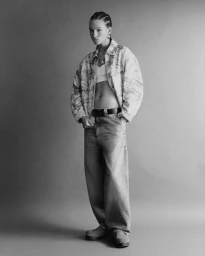 Full-length portrait of a model wearing patterned denim jacket and loose jeans against a plain grey seamless backdrop.