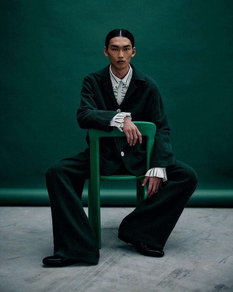 Portrait of Asian male model in a dark green suit sitting on a green chair against a dark green backdrop in a fashion editorial shot.