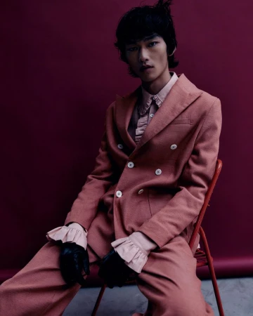 An Asian male model is sitting on a red chair, wearing a rose-colored suit with black gloves in front of a maroon backdrop.