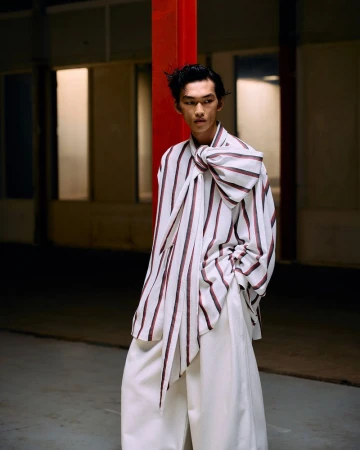 Asian male model wearing a white and striped shirt with a large bow, paired with wide-leg white pants, posing in a dimly lit space.