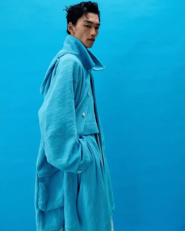 Side view of an Asian male fashion model wearing a textured, light blue jacket against a matching blue backdrop in a studio shot.