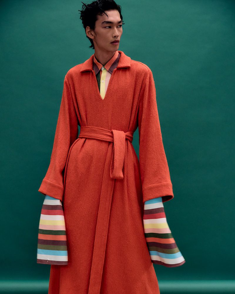 Asian male fashion model poses in a long orange coat with colorful striped sleeves against a solid green backdrop in a studio shot.