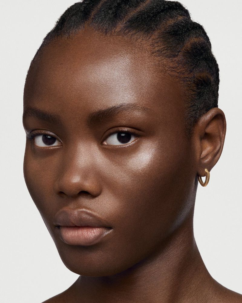 Close-up portrait of a dark-skinned adult woman with cornrow braids, wearing gold hoop earrings against a neutral background.
