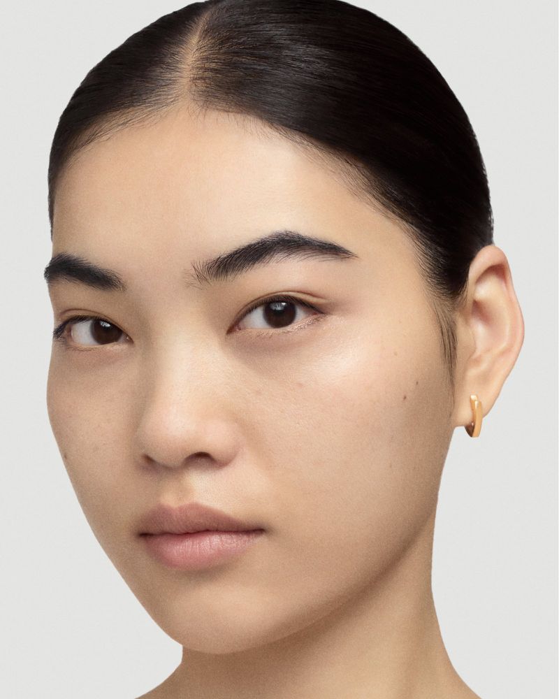 Close-up portrait of a young adult woman with fair skin, dark hair pulled back, wearing small gold hoop earrings against a neutral backdrop.