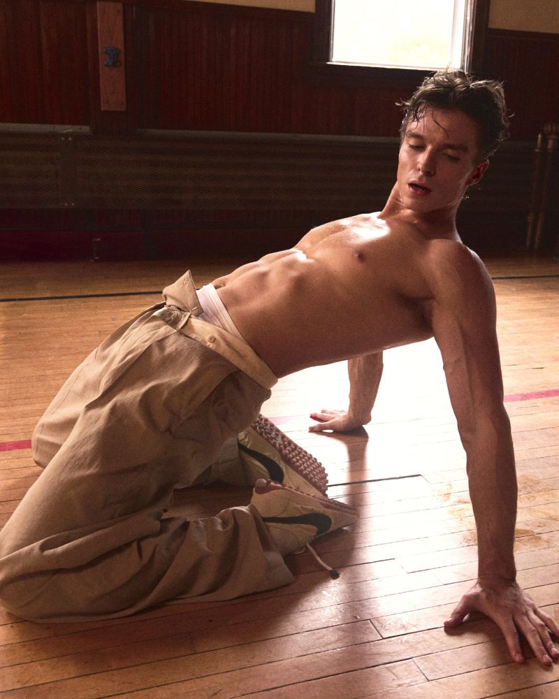 Athletic man in a kneeling backbend with bare torso, wearing tan pants, on a wooden floor in a room with natural light.