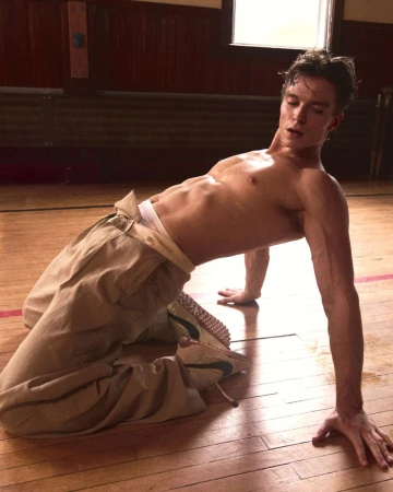 Athletic man in a kneeling backbend with bare torso, wearing tan pants, on a wooden floor in a room with natural light.