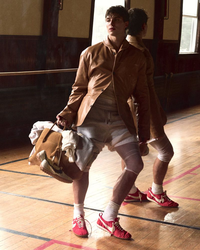 Young adult in a tan jacket and shorts holds a duffel bag while standing in a basketball court, a mirrored reflection behind.