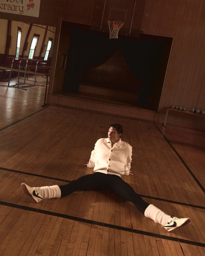 Person sitting on a wooden basketball court in a vintage-style white cardigan, black leggings, and white leg warmers and shoes.