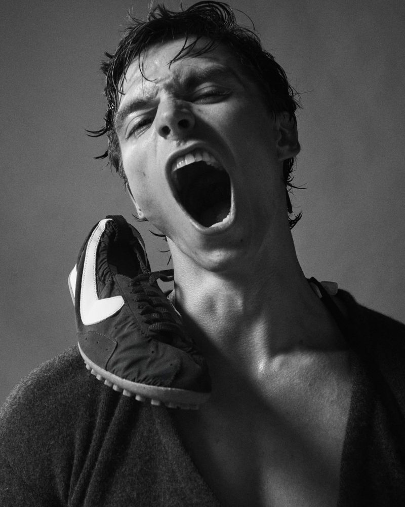 Black and white portrait of a man with an open mouth and a running shoe on his shoulder against a neutral background.