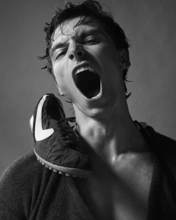 Black and white portrait of a man with an open mouth and a running shoe on his shoulder against a neutral background.