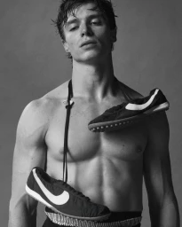 Shirtless man with wet hair posing with sneakers hung around his neck against a plain background
