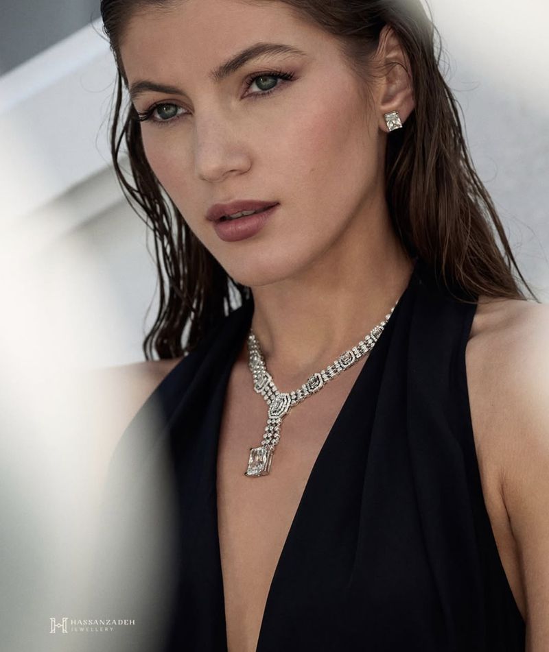 A woman with damp brown hair poses indoors wearing a black top and white gold jewelry necklace and earrings against a blurred background.