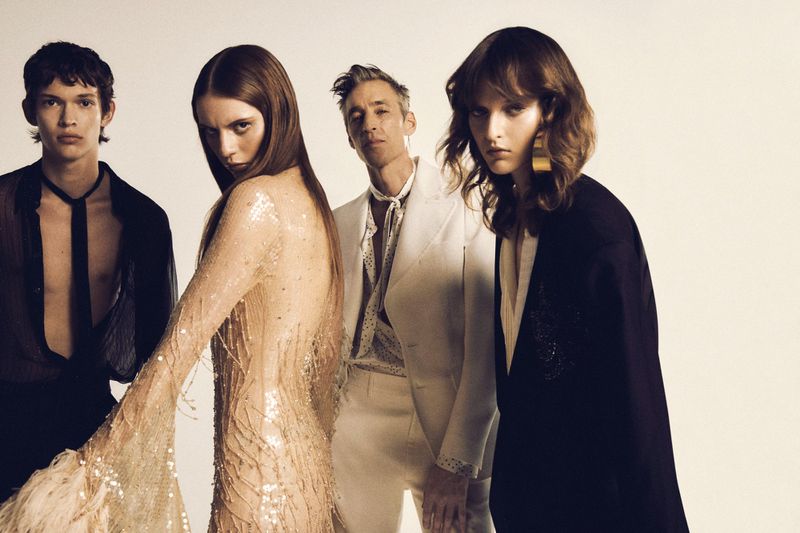 Four fashion models in a studio setting wearing sheer and tailored garments in a group fashion shot, with neutral background.