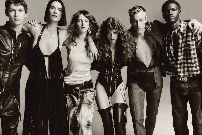 Editorial fashion shot of a diverse group of six young adults with their arms around each other, posing in a studio with a gray background.