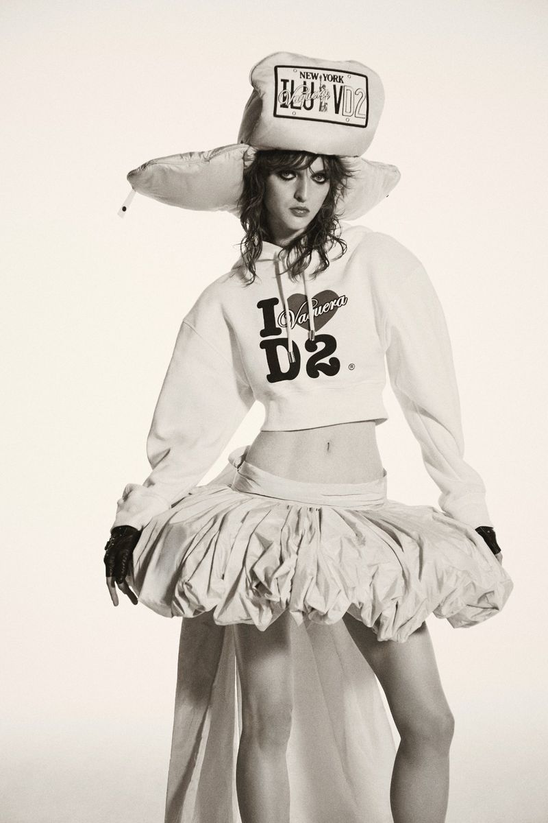 A woman in a white hoodie, tutu skirt, and oversized hat poses in a studio setting with a neutral background.