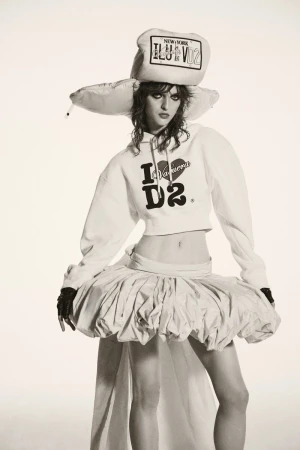 A woman in a white hoodie, tutu skirt, and oversized hat poses in a studio setting with a neutral background.