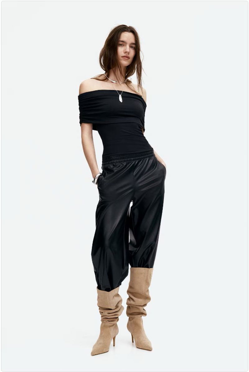 A woman in an off-the-shoulder black top, shiny black pants, and high-heeled boots poses against a white backdrop in a studio setting.