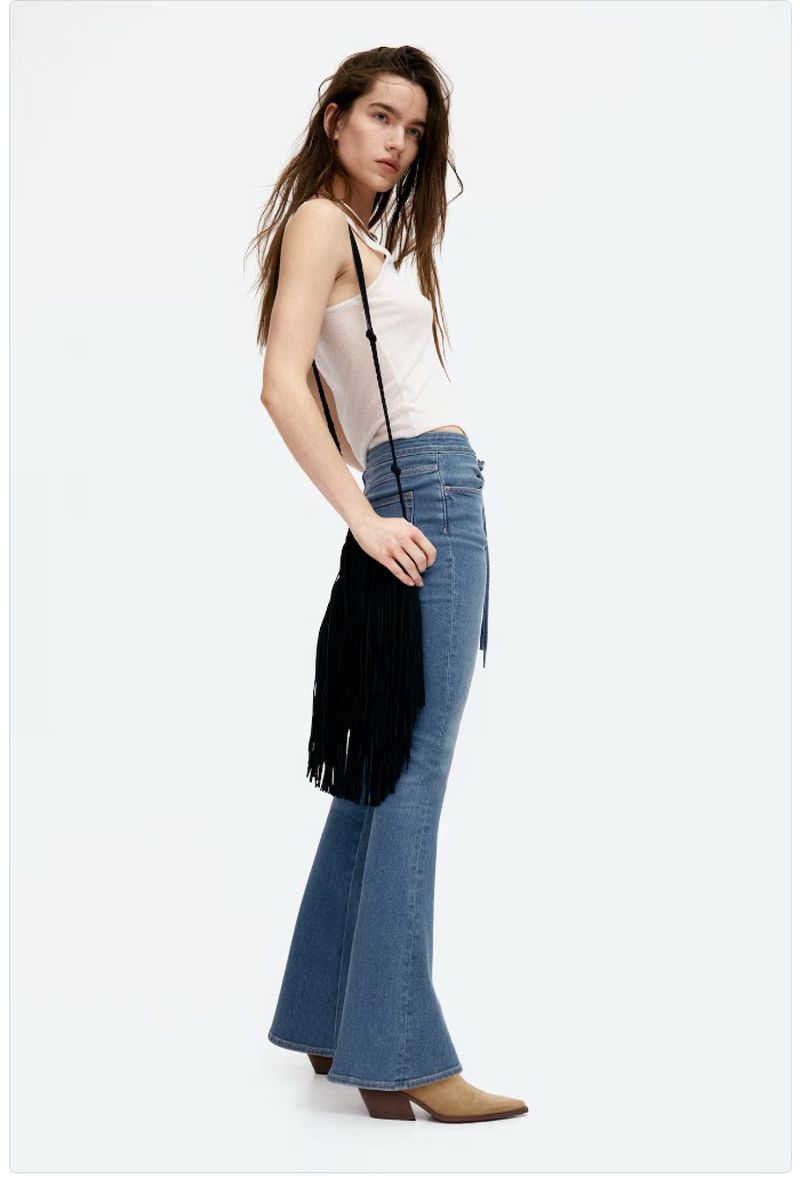 Woman in a white tank top, flared jeans, and heeled booties with a black fringe bag poses in a studio with a white background.