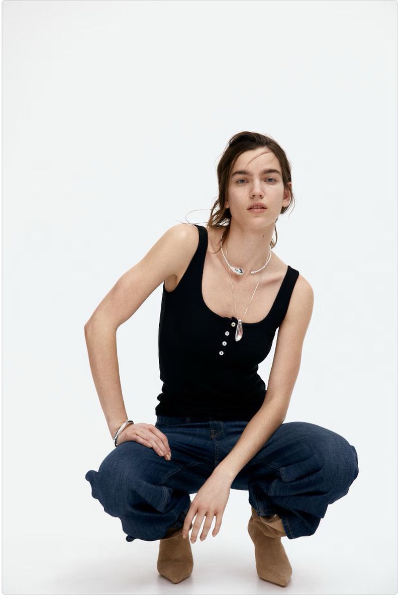 A woman in a black tank top, loose jeans, and tan boots squats on a white background, posing for a fashion or lifestyle shot.