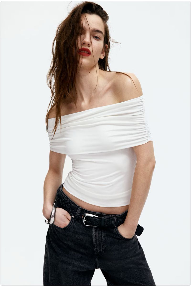 Fashion photo of an adult woman wearing a white, off-the-shoulder top and dark jeans, posing in a photo studio with a white background.