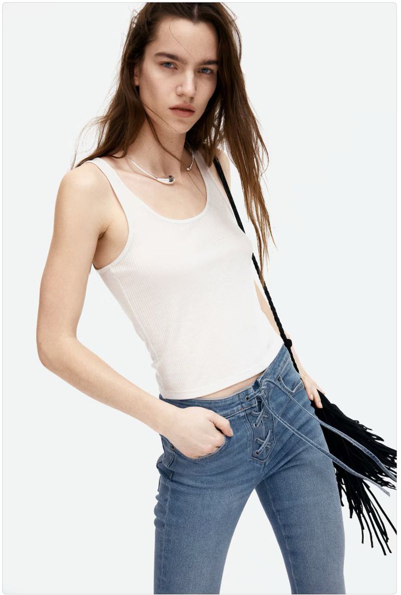 Studio shot of a young adult woman wearing a white tank top and blue jeans, posing against a white background with a handbag.