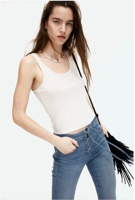 Studio shot of a young adult woman wearing a white tank top and blue jeans, posing against a white background with a handbag.