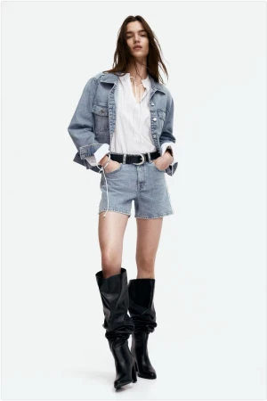 Model in denim jacket and shorts with black boots standing against white seamless background