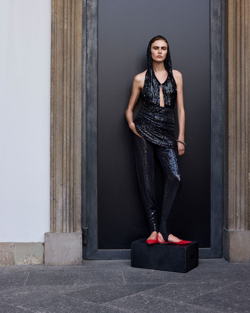 Fashionable woman in a sequined halter top and leggings poses against a dark gray door. Red ballet flats add a pop of color.