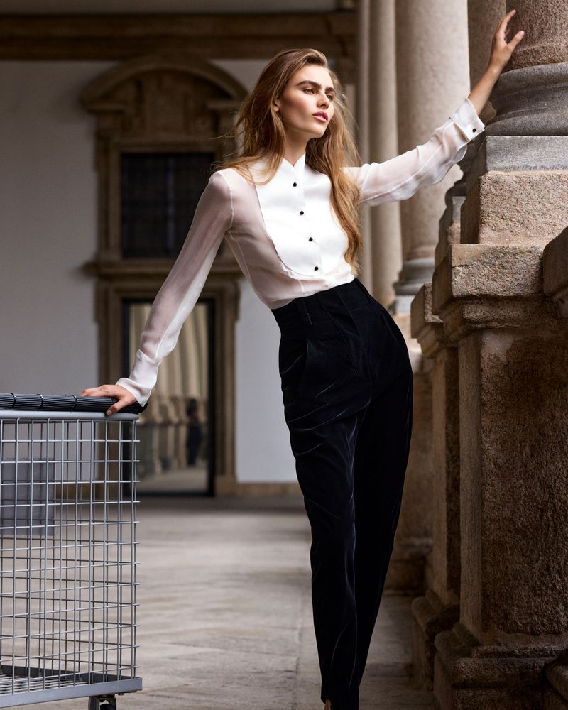 A woman poses elegantly in an architectural setting, wearing a sheer white blouse and dark velvet pants, capturing high fashion.