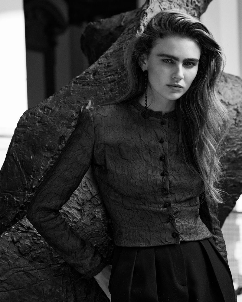 Monochrome portrait of a young woman in a patterned jacket with long wavy hair, posed against a textured sculpture.
