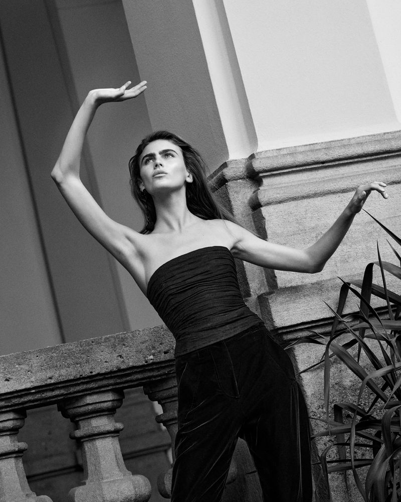 A young adult woman with long dark hair strikes a pose with raised arms on a balcony with foliage in this black and white photograph.
