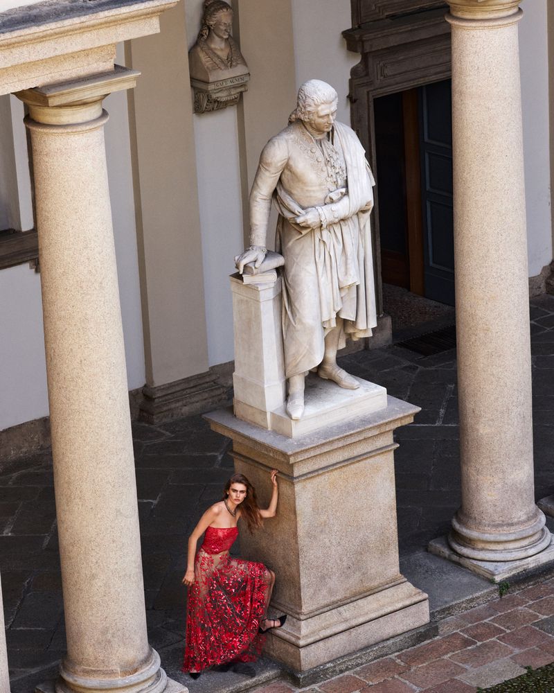 Fashion photo of a woman in a red gown posing next to a marble statue in a courtyard with columns and stone architecture in daytime.