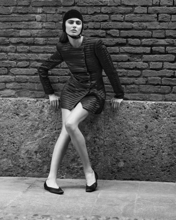 Monochrome fashion portrait of a woman in a patterned mini dress and cap, leaning against a brick wall in an urban setting.