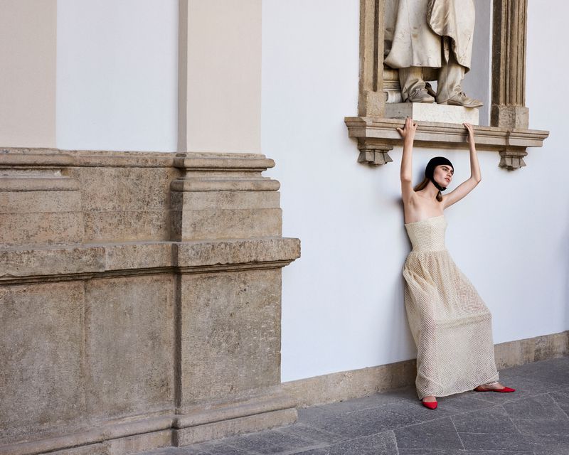 An adult woman in a gown poses outside a building with a stone facade, arms overhead, styled with a black cap and red flats for a fashion shoot.