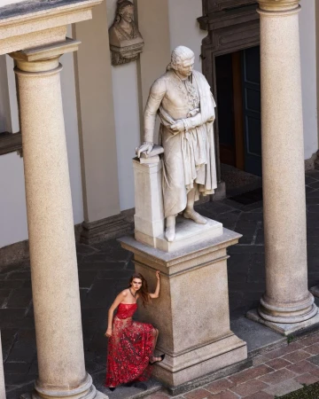 Model Mia Armstrong wearing an embellished red gown leans against a monumental stone statue plinth framed by architectural columns.