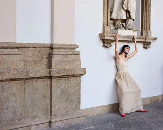 Model Mia Armstrong poses in a strapless cream gown and black headpiece beside a classical stone statue for Vogue Italia.