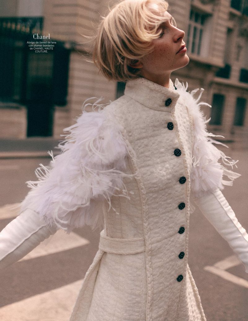 Fashion editorial shot of a woman in a textured white coat with feather epaulettes, outdoors with soft sunlight and blurred street background.